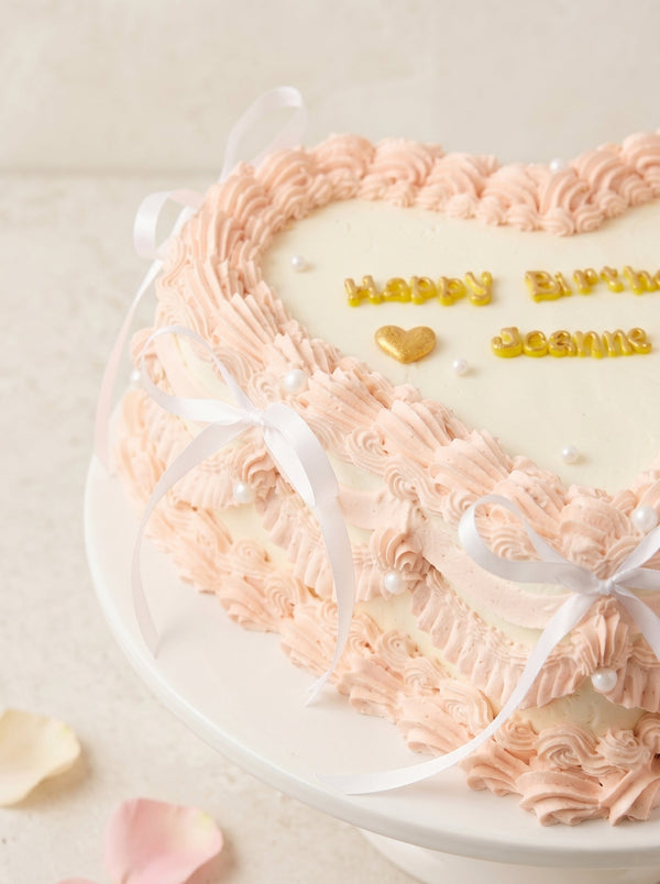 Heart-shaped cake decorated with pale pink piped buttercream, white center panel, pearl decorations, and tied white ribbons.