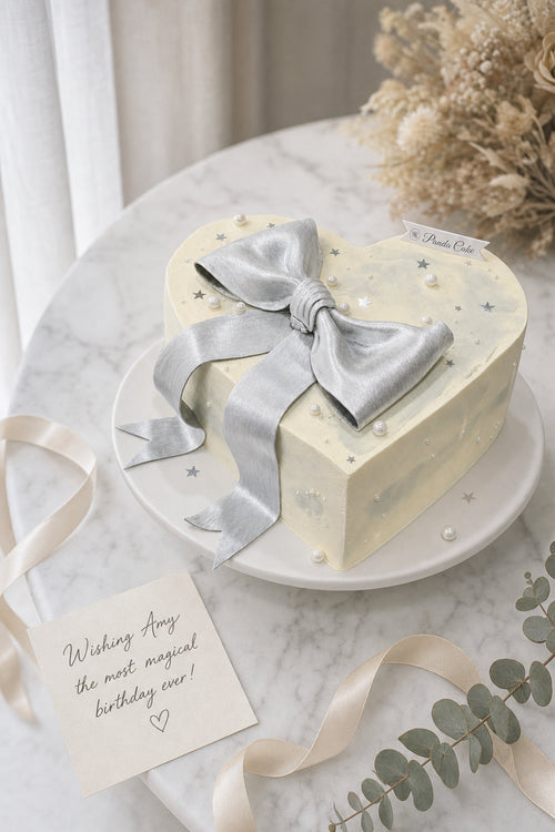 Elegant heart-shaped cream cake featuring a large metallic silver ribbon bow with delicate silver star and pearl decorations