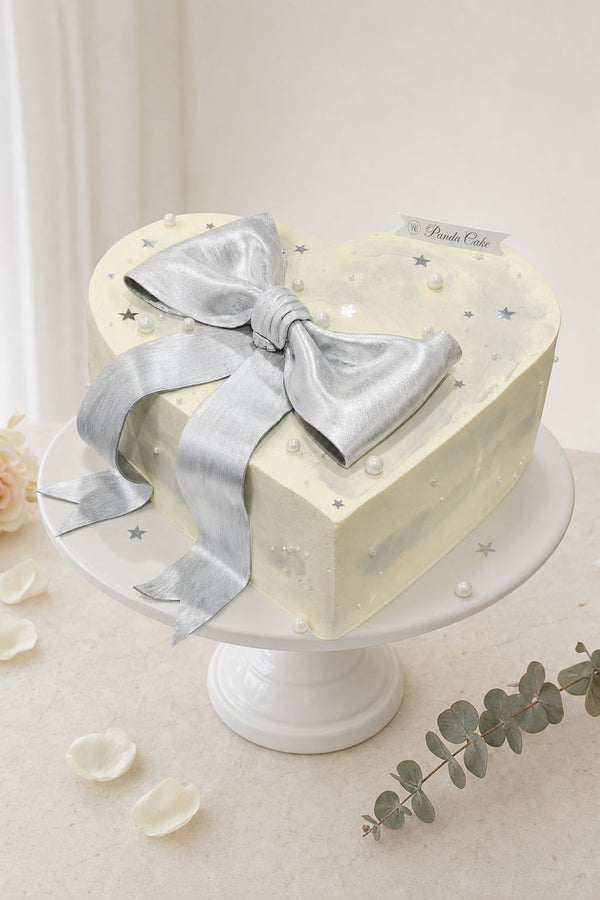 Heart-shaped pale cream cake with large metallic silver ribbon bow and scattered silver stars and pearls