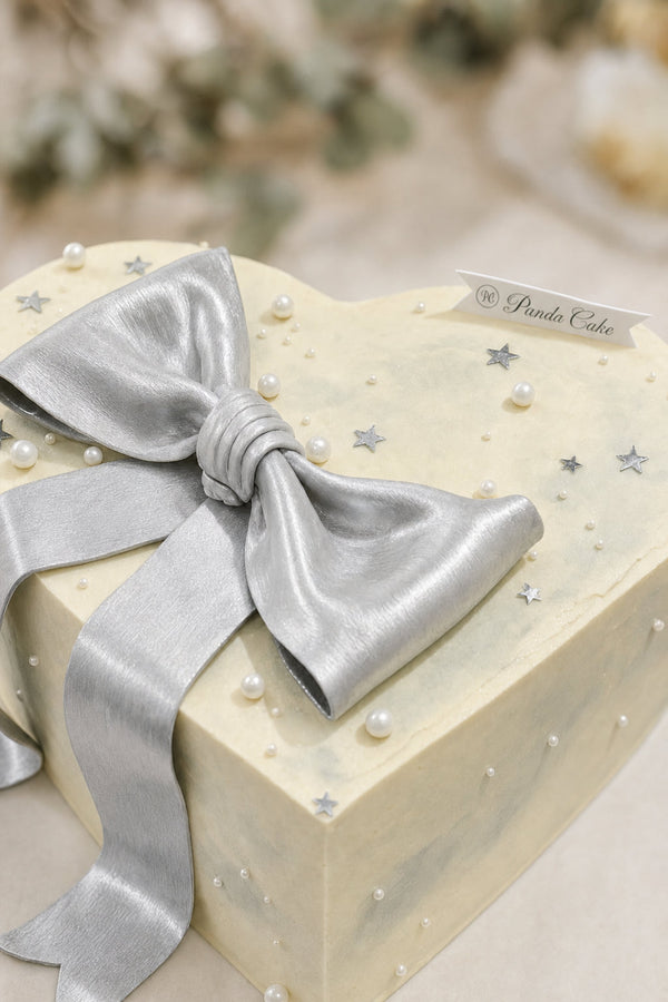 Close-up of heart-shaped cream cake topped with shiny silver bow and decorated with silver star confetti and pearls
