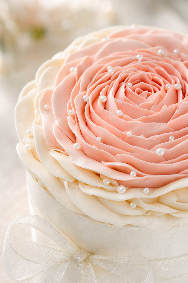 Round white cake topped with a large layered buttercream rose in shades of pink, blush, and ivory, decorated with small pearl accents