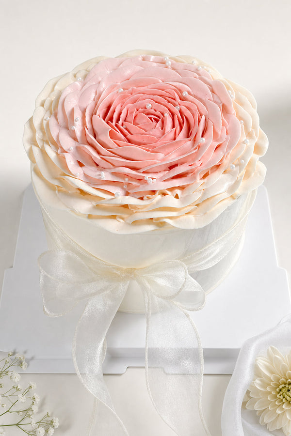 Studio product hero image of an elegant round birthday cake featuring a large pink to blush buttercream rose on top with a white ribbon tied around the base