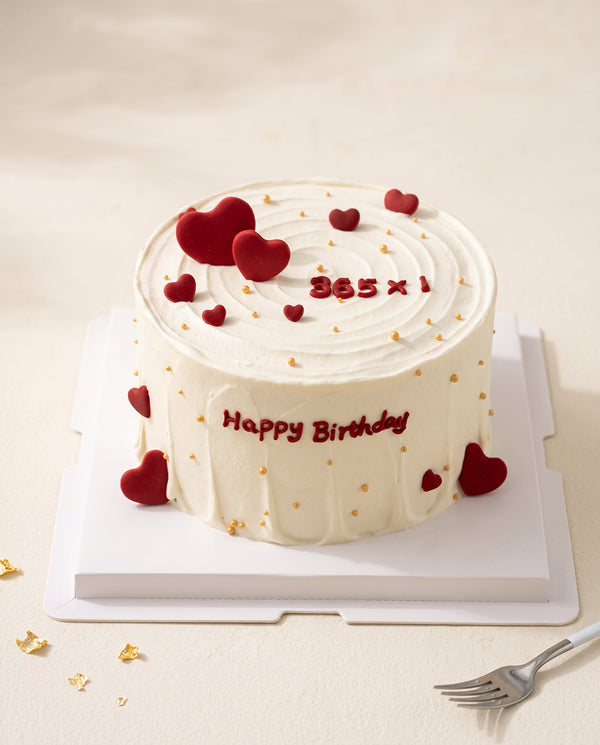 Round white buttercream birthday cake with smooth spiral top, red heart toppers and gold pearl sprinkles on lace background