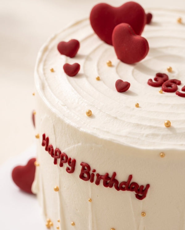 Close-up of elegant round birthday cake, white buttercream spiral finish with small red hearts and golden sprinkles
