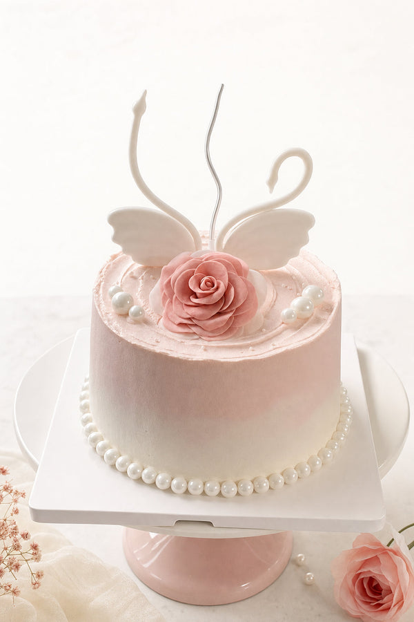 Studio hero photograph of round pale pink buttercream cake with blush pink fondant rose and white swan-shaped topper with curved necks