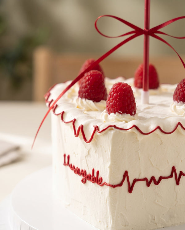 White buttercream round cake decorated with red scalloped piping and fresh raspberries on top, held by a person wearing black.