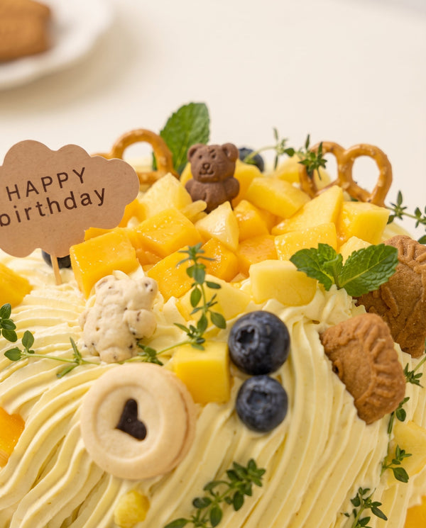Square cream-frosted birthday cake with piped ridges topped with fresh mango cubes, blueberries, cookies, pretzels and mint leaves