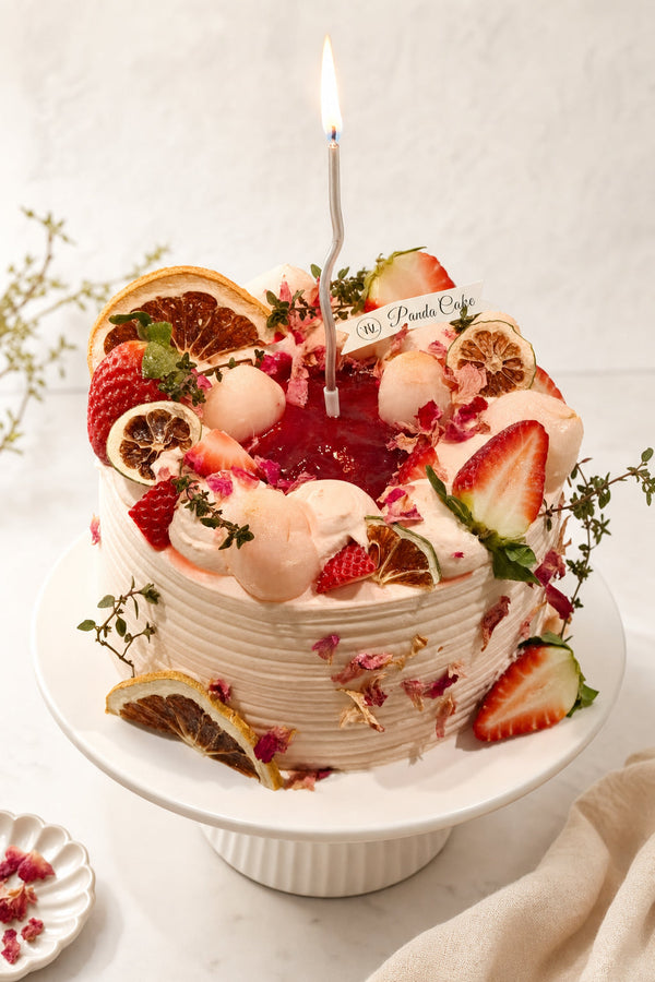 Studio hero shot of a white frosted birthday cake with glossy red center topped with fresh strawberries, dried citrus slices, rose petals and cream dollops