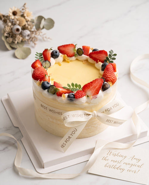 Tall round cream cake with smooth pale yellow frosting, topped with halved strawberries and blueberries
