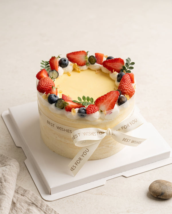 Studio hero image of a tall round pale yellow cream cake with neat cream ring and fresh strawberry and blueberry crown