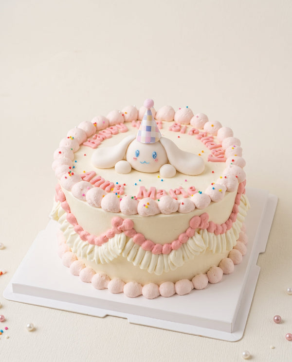 Close-up studio image of a round pastel cream cake with pink and white piped border and a bunny figure wearing a party hat on top