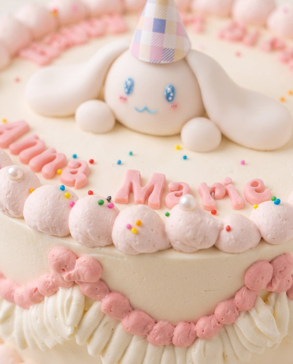 Round celebration cake with soft pastel cream base, pink piped borders, pearl-like dollops, and multicolor sprinkles