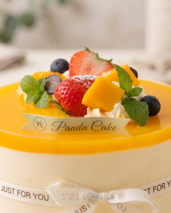 Glossy mango mousse cake with orange glaze, whipped cream, sliced strawberries, blueberries, and mango cubes