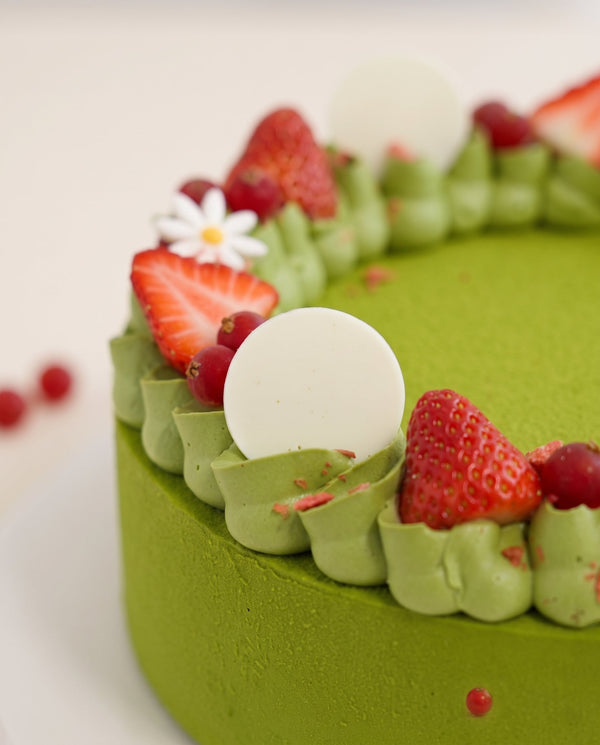 Close-up of a green mousse cake decorated with strawberries, raspberries, white chocolate discs and edible flowers on a white cake board
